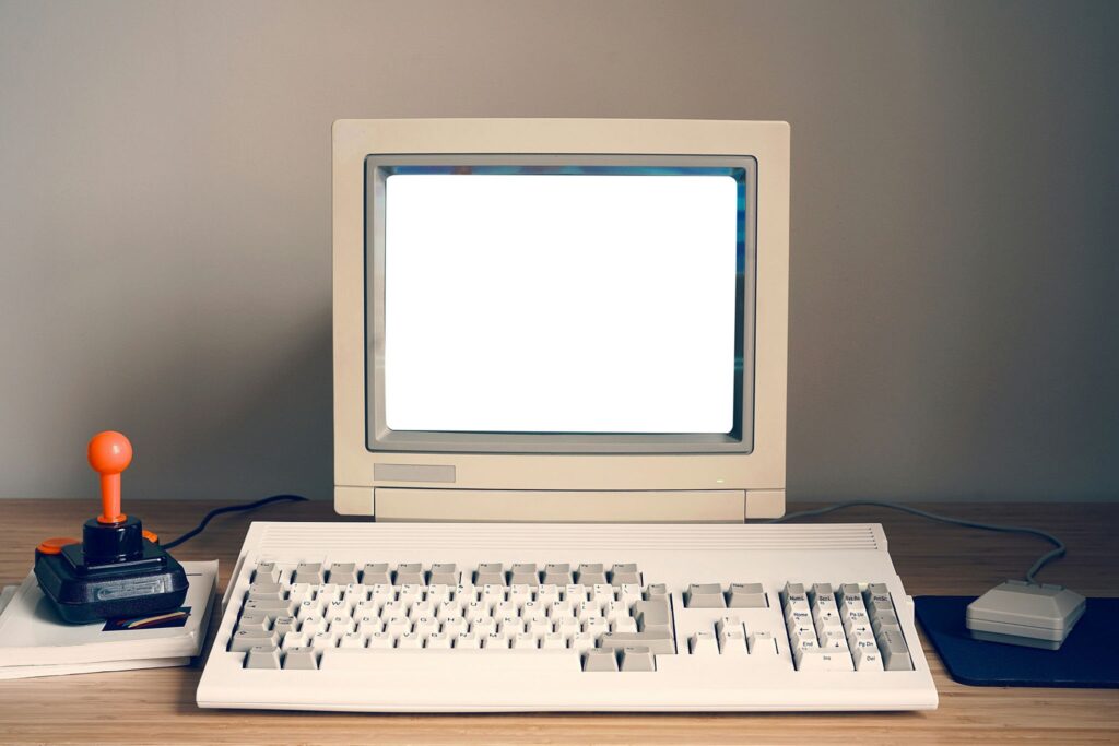 Fotografía de un escritorio con un ordenador retro de finales de los años 80 y principios de los 90. En el centro destaca un monitor de tubo CRT de color beige encendido con la pantalla en blanco, un teclado mecánico de perfil alto sobre la mesa, un ratón antiguo sobre una alfombrilla azul a la derecha y un joystick clásico de palanca con bola naranja a la izquierda
